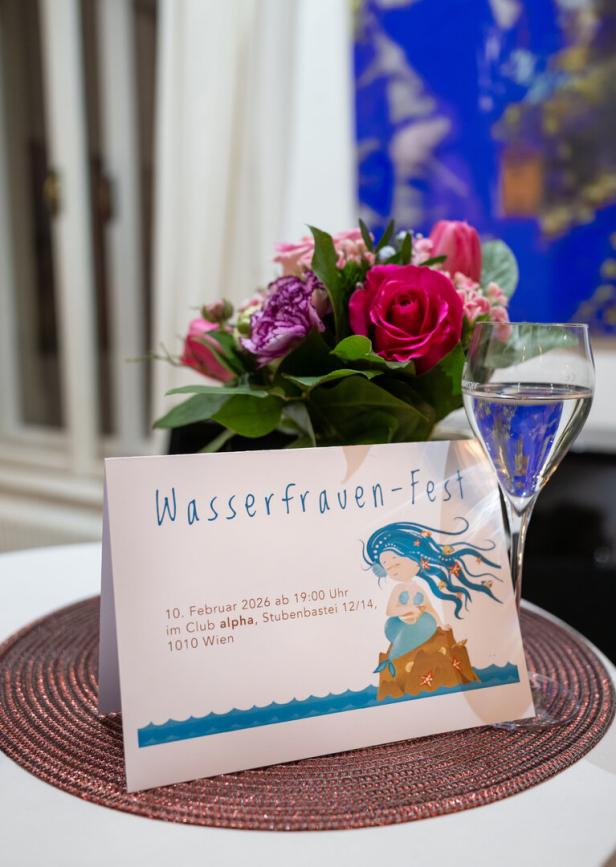 Ein Strauß aus rosa und lila Blumen, ein Glas Wasser und eine Einladung zum Wasserfrauen-Fest auf einem Tisch.