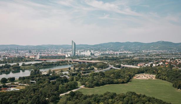 Grüne Parklandschaft mit Fluss, mehreren Brücken, moderner Skyline und Hügeln im Hintergrund unter leicht bewölktem Himmel.