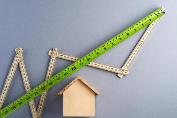 Ein kleines Holzhaus und aufgeklappte Zollstöcke symbolisieren die Wertentwicklung und Planungssicherheit von Immobilieninvestitionen.