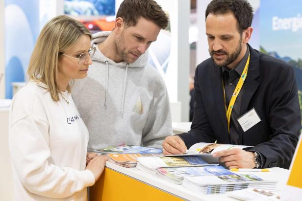 Drei Personen im Beratungsgespräch an einem Messestand mit Broschüren auf dem Tresen.