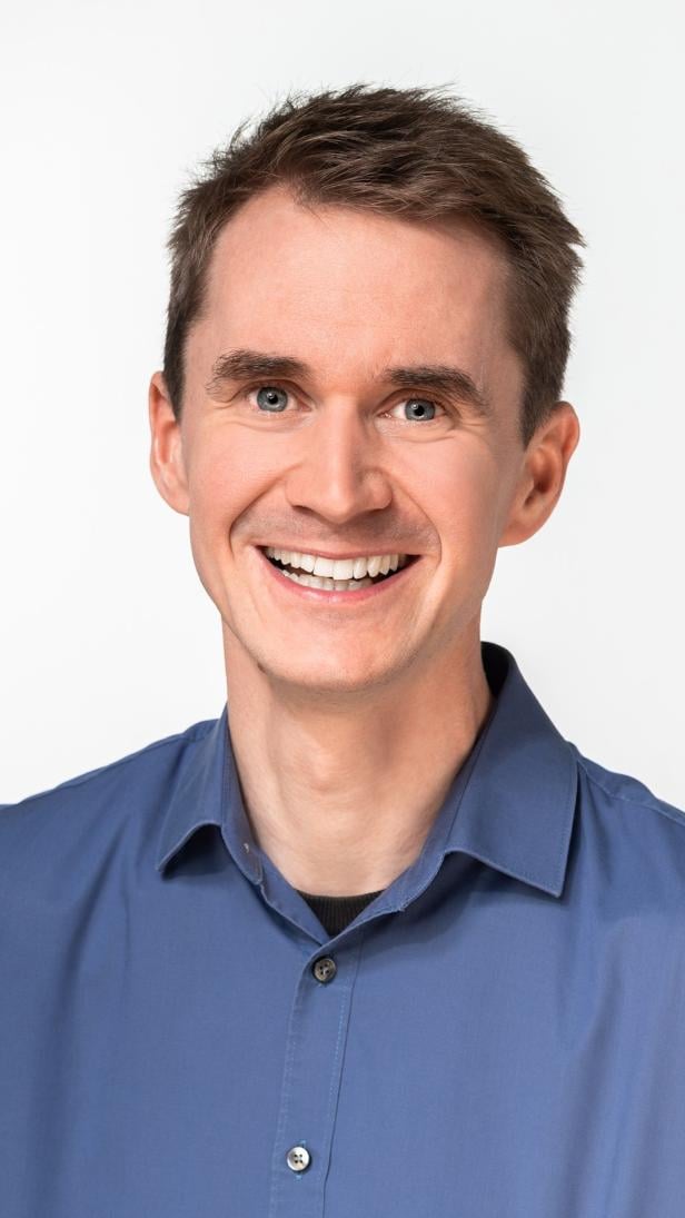 Mann mit kurzem braunem Haar und blauem Hemd lächelt vor hellem Hintergrund.