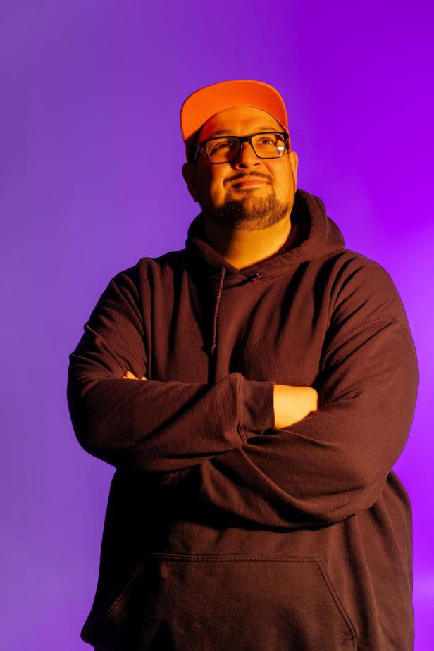 Mann mit Brille und orangener Kappe vor lila Hintergrund, verschränkt die Arme und lächelt.