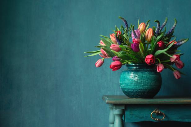 Ein Blumenstrauß mit Tulpen und blauen Blüten steht in einer türkisfarbenen Vase auf einem Tisch.