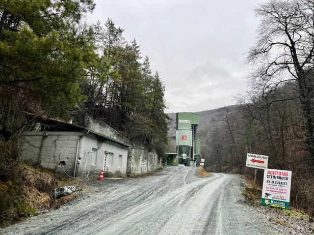 Eine Schotterstraße führt zu einer Industrieanlage im Wald, mit Warnschildern und einem alten Gebäude am Straßenrand.