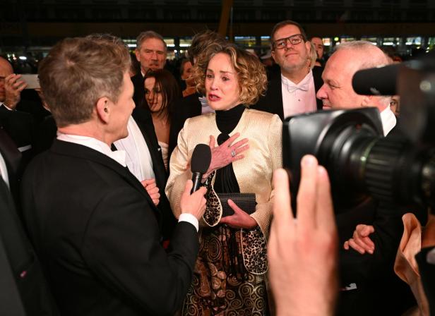 Eine elegant gekleidete Frau wird auf einem roten Teppich von einem Reporter interviewt, umgeben von mehreren Menschen.