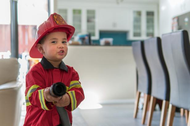Ein Kind in Feuerwehrkostüm hält einen Schlauch und steht in einem modernen Wohnzimmer.
