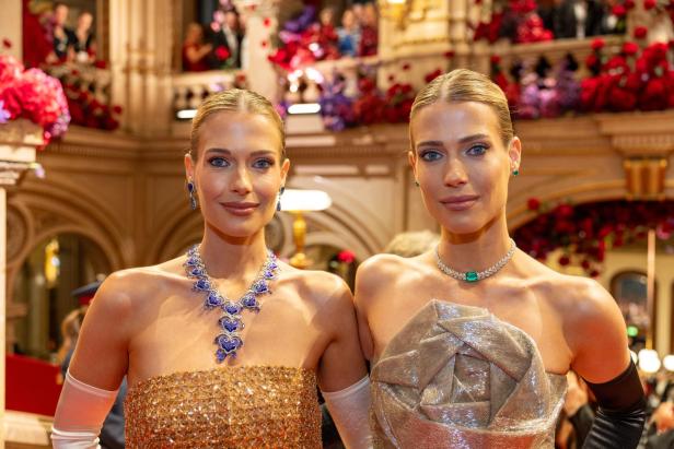 Zwei elegant gekleidete Frauen mit auffälligem Schmuck posieren in einem festlich geschmückten Saal.