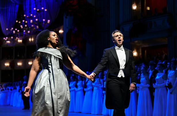 Ein elegant gekleidetes Paar singt auf einer festlich beleuchteten Bühne und hält sich an den Händen.