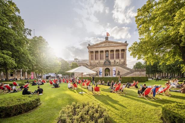 Menschen sitzen entspannt auf Liegestühlen und auf dem Rasen vor einem historischen Gebäude im Sonnenschein.
