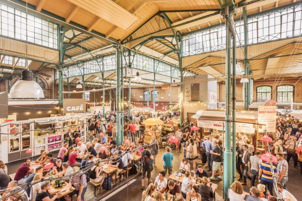 Viele Menschen sitzen und stehen an Tischen und Essensständen in einer hellen Markthalle mit hohen Fenstern.