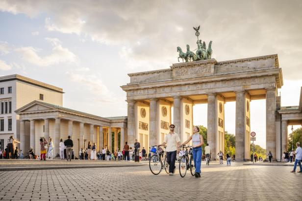Zwei Menschen schieben Fahrräder vor dem Brandenburger Tor, umgeben von vielen weiteren Passanten.