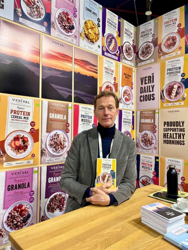 Ein Mann sitzt an einem Tisch und hält eine Packung Protein-Porridge vor einer Wand mit Frühstücksprodukten von Verival.