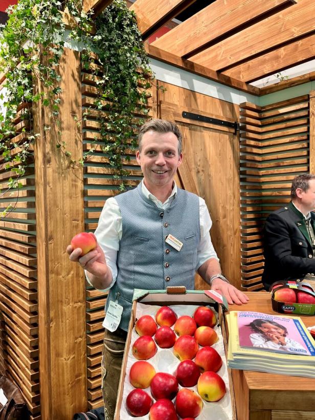 Ein Mann in Tracht hält einen Apfel und steht hinter einer Kiste mit roten Äpfeln an einem Holzstand.