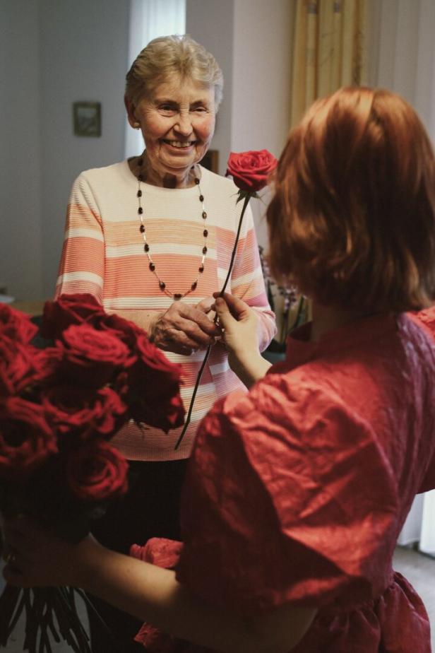 Eine ältere Frau lächelt, während sie von einer jüngeren Person eine rote Rose überreicht bekommt.