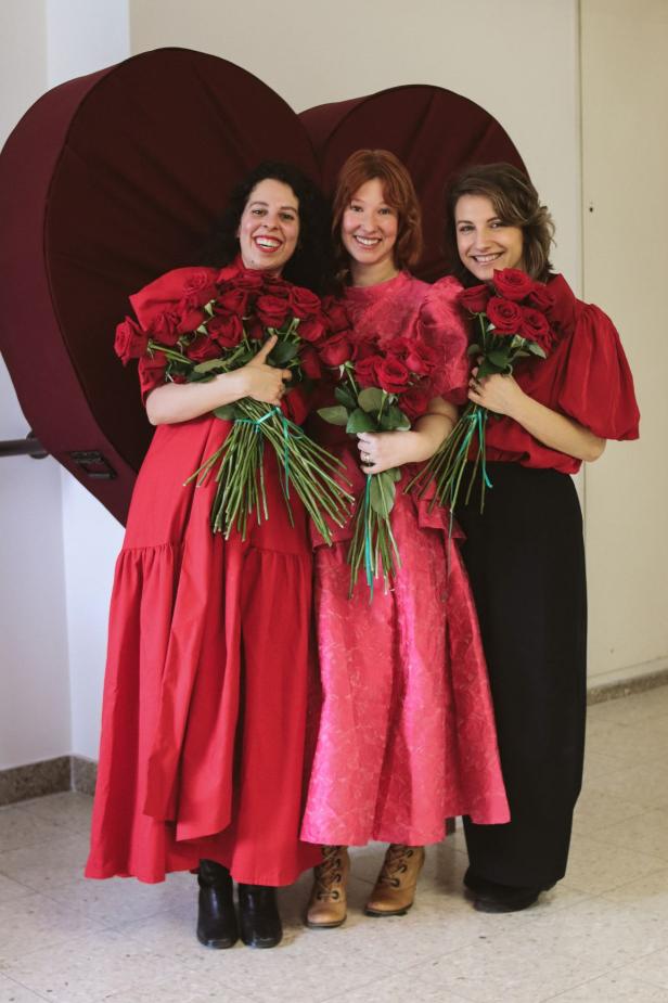 Drei Frauen in roten Kleidern mit Rosen vor einem großen Herz