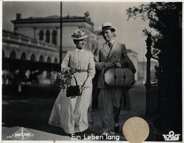 Ein Paar in Hüten und Kleidung der 1930er Jahre geht mit einem Koffer auf der Straße.