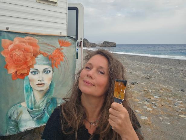 Ein Foto von Doris Breuer, die einen Pinsel in der Hand hält und in die Kamera schmunzelt. Hinter ihr ist ein Gemälde von einer Frau in blau und orange Tönen.