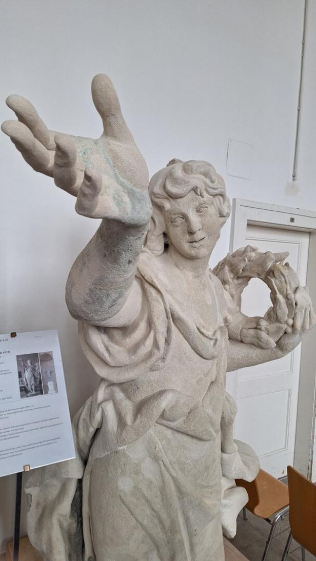 Die Statue eines steinernen Engels, der in einer Hand einen Kranz hält und die andere in Richtung Himmel streckt.