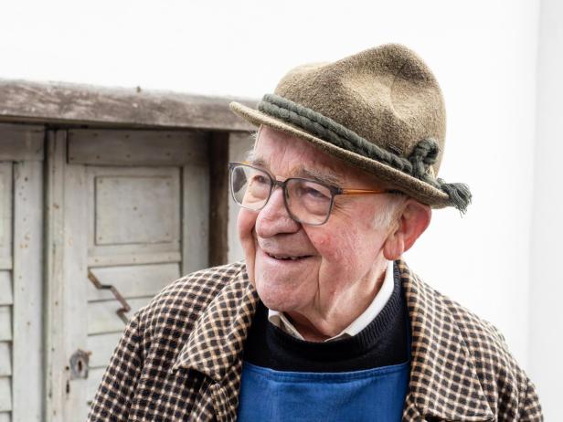 Ein älterer Mann mit Brille und Trachtenhut lächelt, trägt karierten Mantel und steht vor einer alten Holztür.