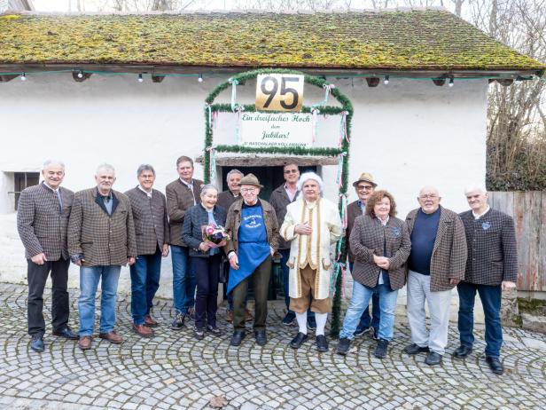 Vierzehn ältere Menschen stehen lächelnd vor einem kleinen, weiß getünchten Haus mit Moosdach und einer „95“-Jubiläumsdekoration.