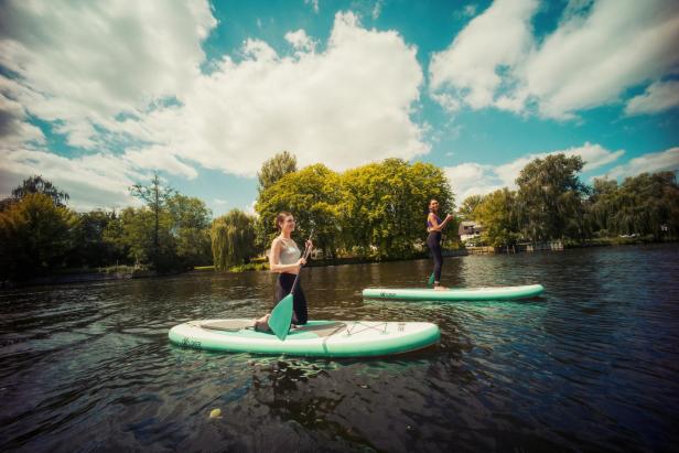 Zwei Frauen paddeln auf SUP-Boards auf einem ruhigen Fluss, umgeben von grünen Bäumen und blauem Himmel.