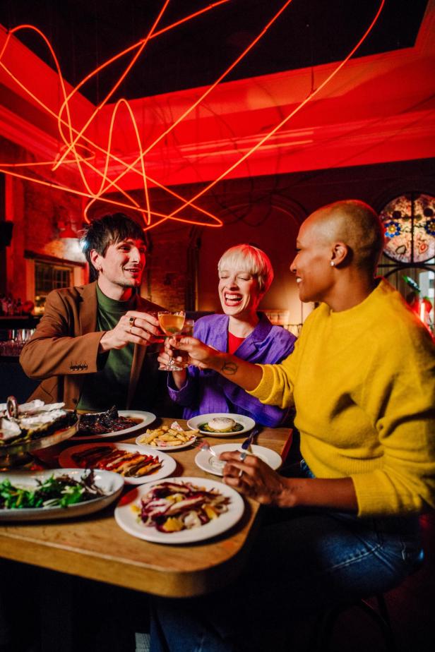 Drei Menschen stoßen lachend mit Getränken an einem Tisch voller Speisen in einem stimmungsvoll beleuchteten Restaurant an.