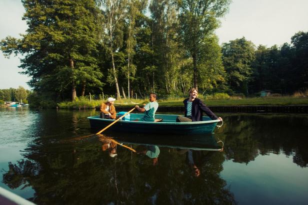 Drei Personen sitzen in einem Ruderboot auf einem ruhigen See, umgeben von grünen Bäumen.