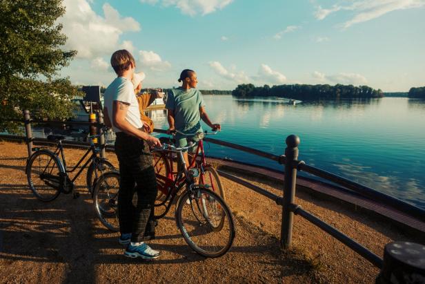 Drei Personen mit Fahrrädern stehen an einem Seeufer und blicken gemeinsam auf das Wasser.
