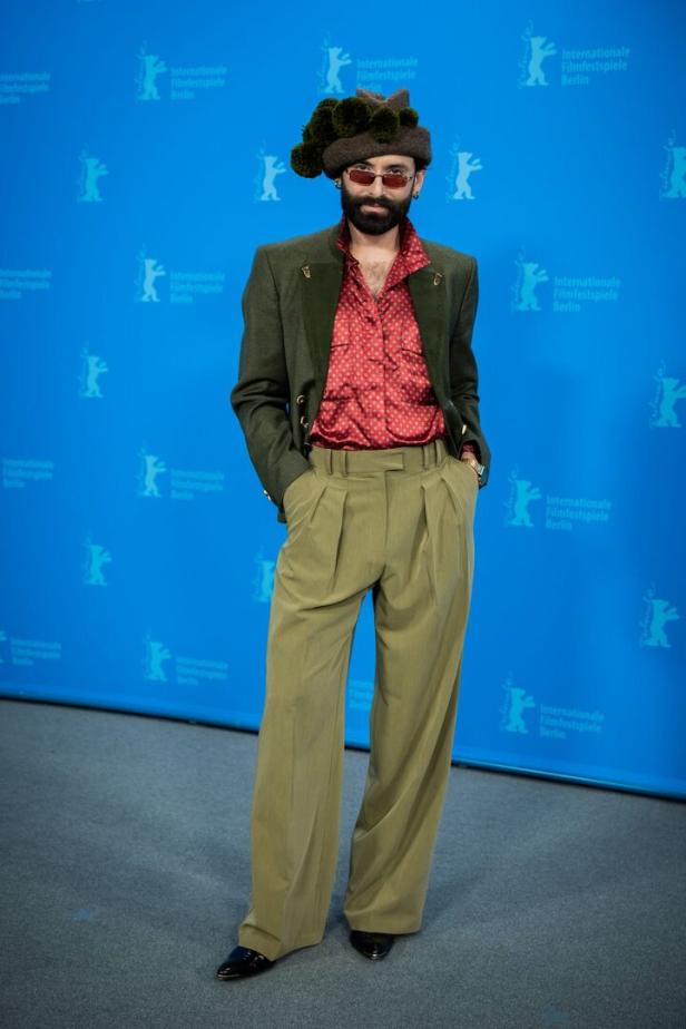 Eine Person mit auffälligem Hut, rotem Hemd, grünem Blazer und weiter Hose steht vor blauem Berlinale-Hintergrund.