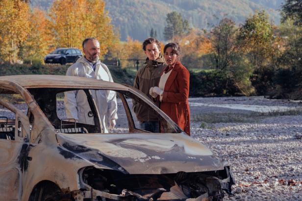 Filmszene: Drei Personen stehen vor einem ausgebrannten Auto in einer Flusslandschaft.