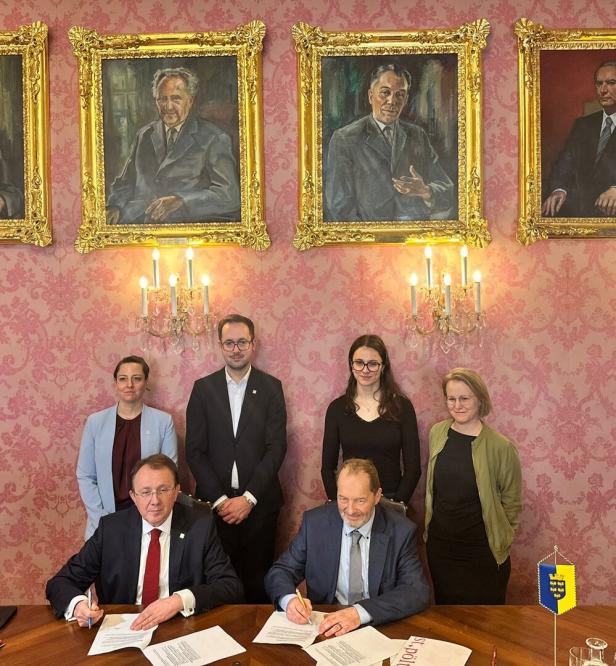 Zwei Personen sitzen an einem Tisch, vier stehen dahinter.