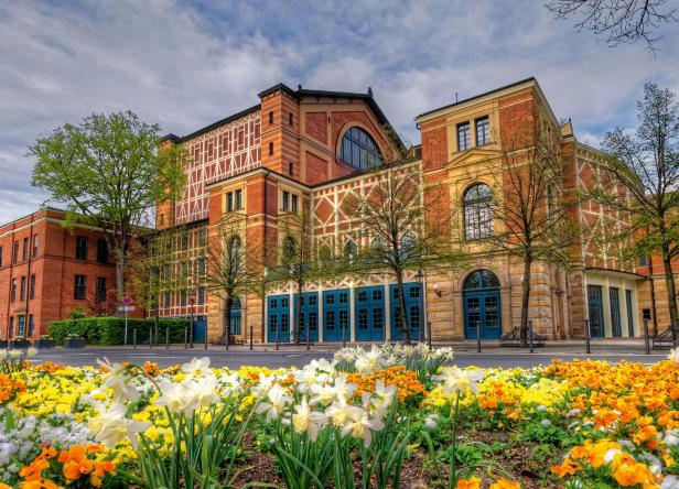 Festspielhaus Bayreuth mit blühendem Blumenbeet im Vordergrund und Bäumen davor.