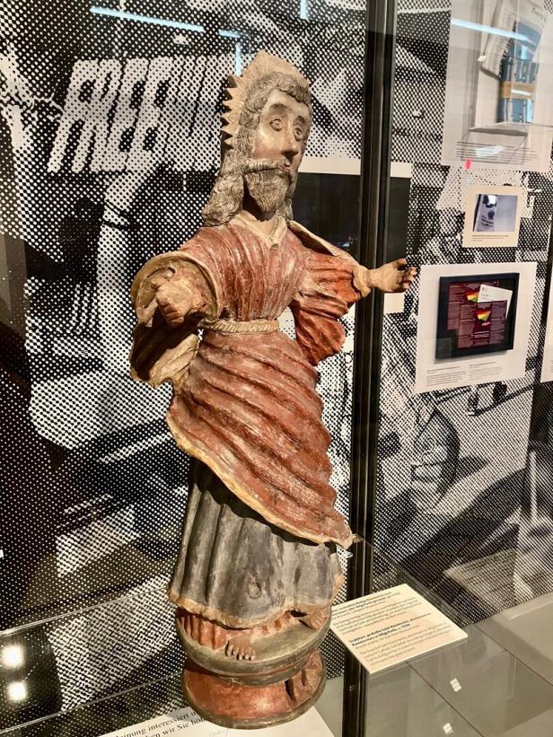 Holzskulptur einer bärtigen Figur mit ausgestreckten Armen, gekleidet in ein rotes Gewand mit grauem Unterrock.