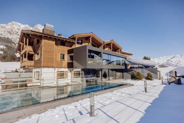 Ein Holzhaus mit Pool liegt in einer verschneiten Winterlandschaft vor einem blauen Himmel.