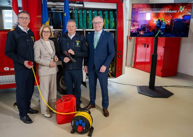 Vier Personen stehen vor einem Feuerwehrfahrzeug mit Ausrüstung, daneben ein Bildschirm mit Feuerwehrmotiv und Schriftzug.