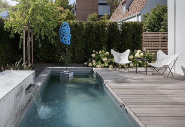 Moderner Garten mit Pool, Holzterrasse, zwei weißen Stühlen und blauer Skulptur.