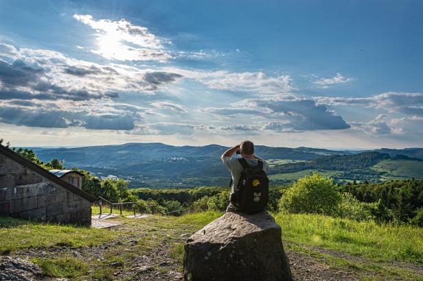 Ein Wanderer mit Rucksack genießt den weiten Ausblick über die hügelige Landschaft der Rhön am Hochrhöner bei Sonnenschein.