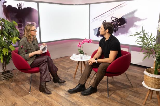 Zwei Personen sitzen sich in roten Sesseln gegenüber und unterhalten sich in einem modern eingerichteten Studio.