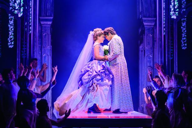 Ein Brautpaar steht sich auf einer Bühne gegenüber, umgeben von jubelnden Menschen in festlicher Kulisse.