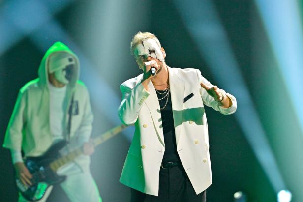 Ein Sänger mit weißer, rissiger Maske und weißem Sakko performt auf einer Bühne, daneben ein Musiker mit Kapuze und Gitarre.