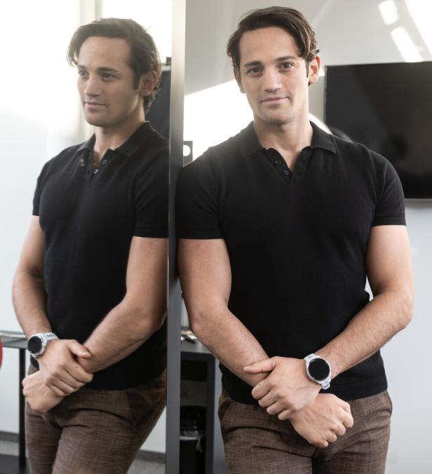 Ein Mann steht vor einem Spiegel, trägt ein schwarzes Poloshirt und eine Uhr, sein Spiegelbild ist deutlich zu sehen.