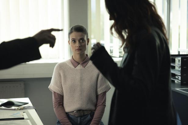 Daphne Meindl (Philine Schmölzer) sitzt ernst zwischen zwei streitenden Personen in einem hellen Raum mit Jalousien.