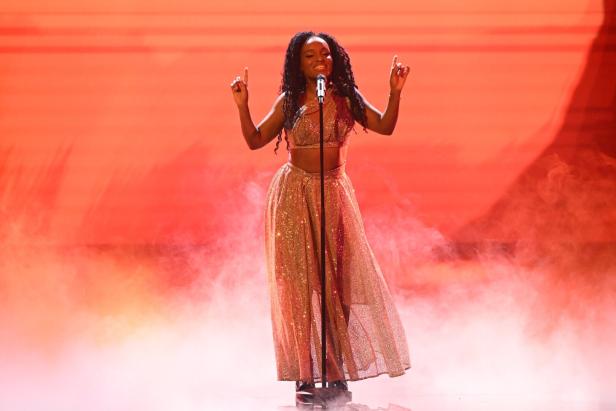 Frau mit langen Locken singt in glitzerndem Outfit auf einer Bühne mit rotem Hintergrund und Nebel.