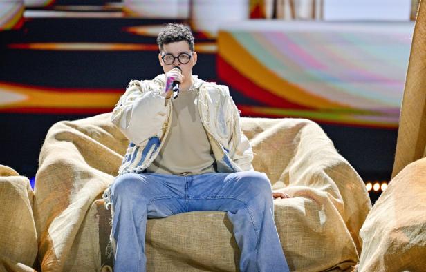 Eine Person mit Brille sitzt auf einem großen, mit Stoff bedeckten Sessel und singt in ein Mikrofon.