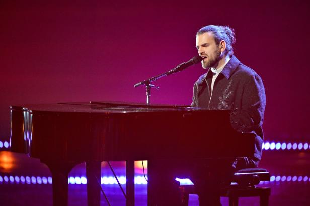 Ein Mann spielt Klavier und singt in ein Mikrofon auf einer Bühne mit lila Hintergrundbeleuchtung.