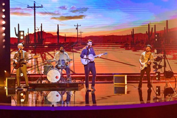 Vierköpfige Band spielt auf einer Bühne mit Wüstenlandschaft und Sonnenuntergang im Hintergrund.