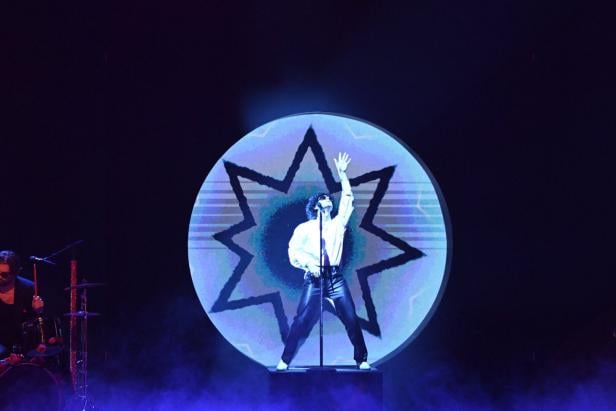 Eine Person steht auf einer Bühne vor einem leuchtenden Kreis mit Sternmuster und hebt eine Hand in die Luft.