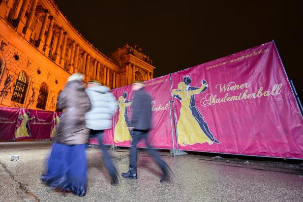 Mehrere Personen gehen an Absperrungen mit der Aufschrift „Wiener Akademikerball“ vor einem beleuchteten historischen Gebäude vorbei.