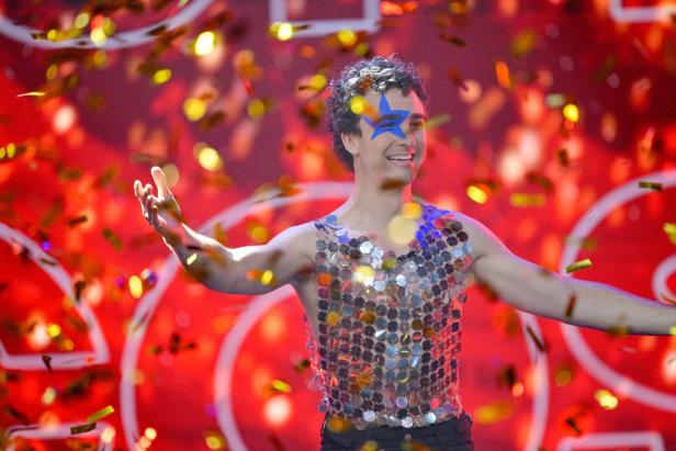 Cosmó jubelt mit ausgebreiteten Armen und blauem Stern im Gesicht auf der Bühne, während goldene Konfetti durch die Luft fliegen.
