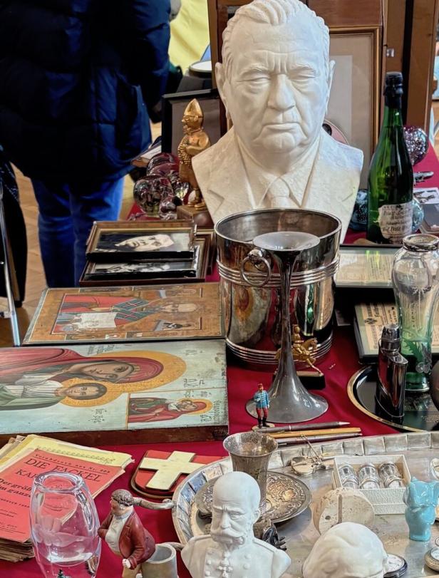 Verschiedene Antiquitäten wie Büsten, Ikonen, Silbergeschirr, Figuren und alte Bücher liegen auf einem Tisch ausgebreitet.